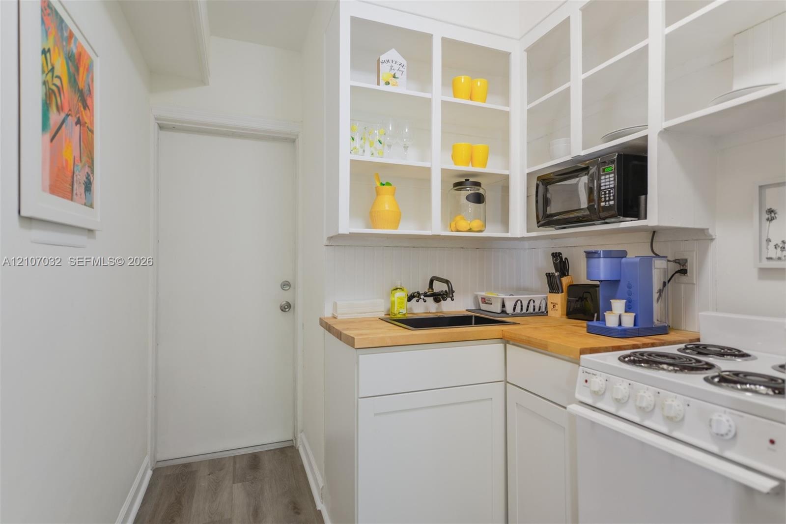 Kitchenette featuring white shaker-style cabinetry, open shelving with glass-front uppers, butcher block countertops, a black undermount sink with a m
