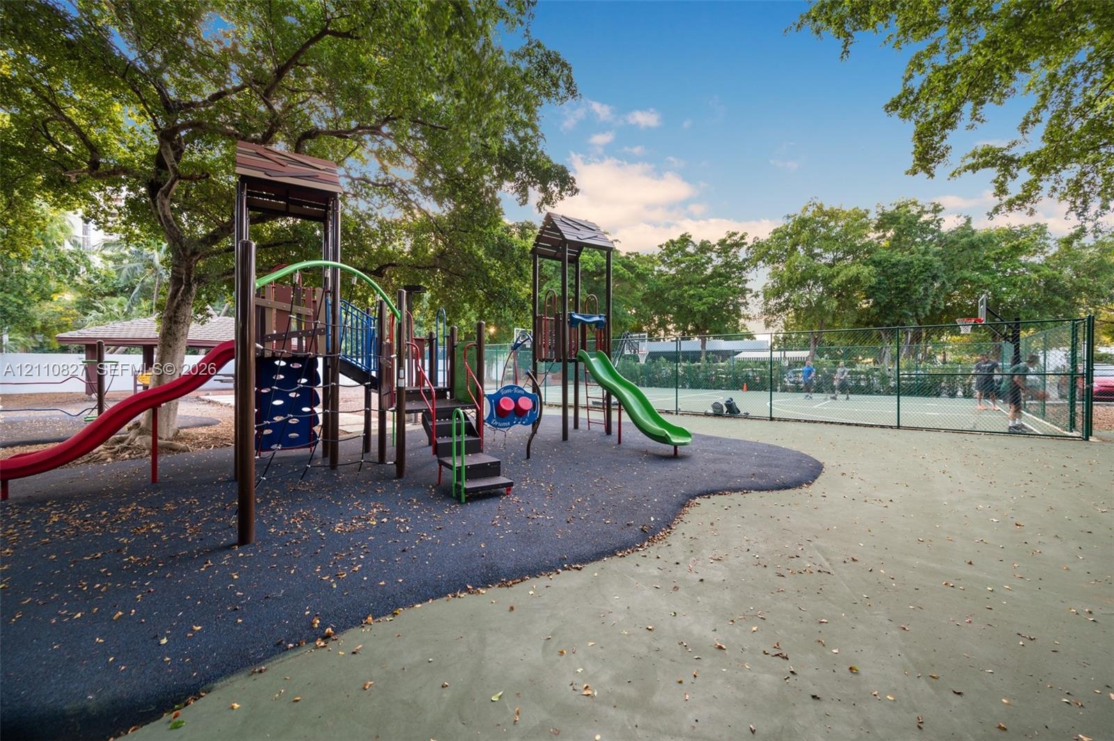 Recreational area featuring a multi-component playground structure, paved sport court, fenced perimeter, and mature canopy trees