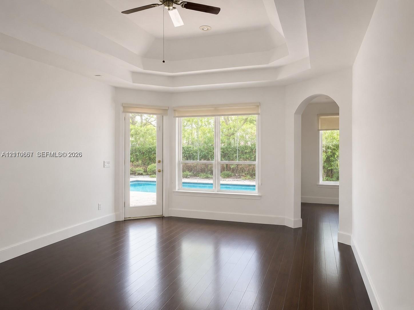 Tray ceiling, wood-finish flooring, and arched entryway featuring large windows and an exterior door providing visibility to the pool area