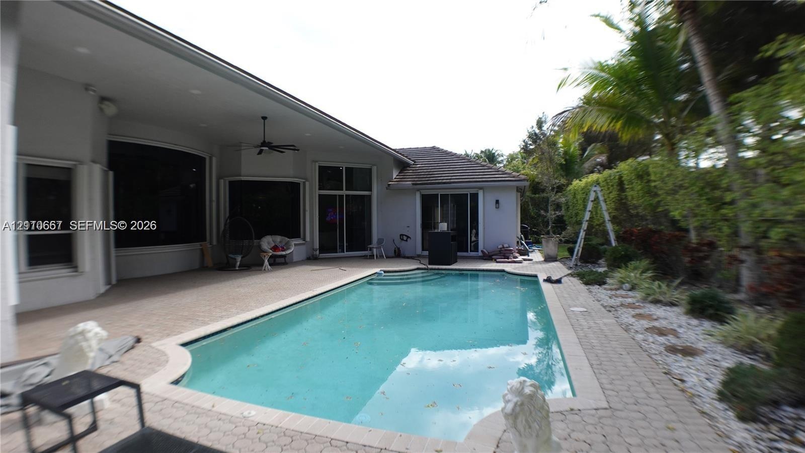 Covered patio with integrated ceiling fan, in-ground pool with stone paver decking, and mature tropical landscaping