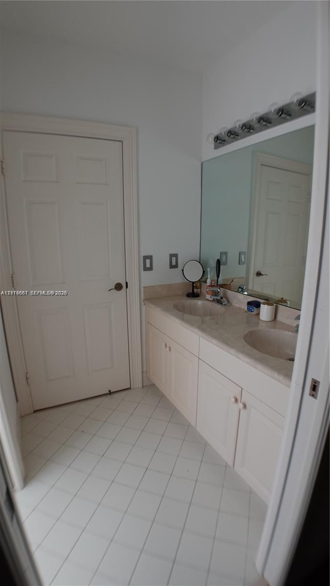 Double vanity featuring integrated sinks, stone-surface countertop, white cabinetry, large wall-mounted mirror, and tile flooring laid on a diagonal