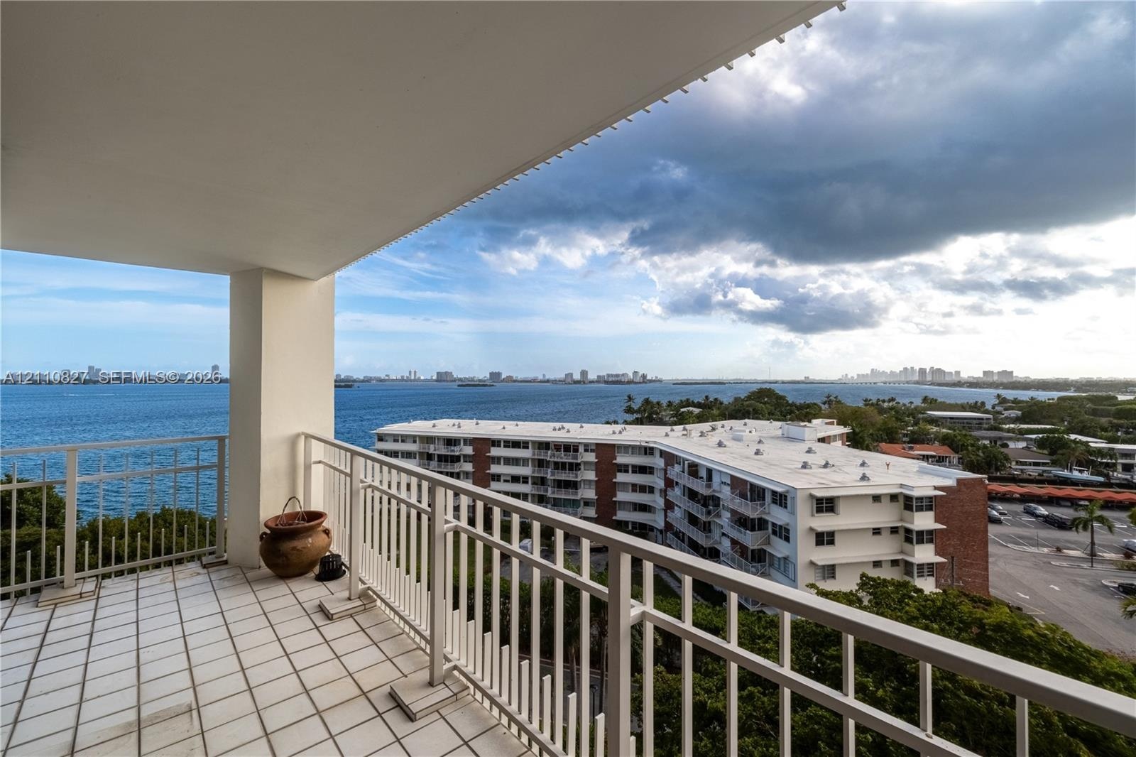 Covered balcony featuring white tiled flooring and a metal railing, overlooking the water and shoreline
