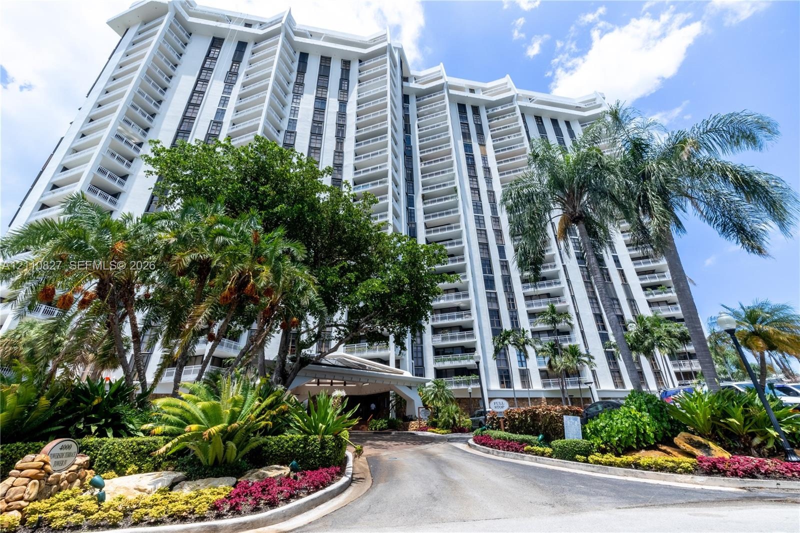 High-rise residential exterior featuring stepped architectural balconies, white concrete facade, covered porte-cochere entry, and extensive tropical l