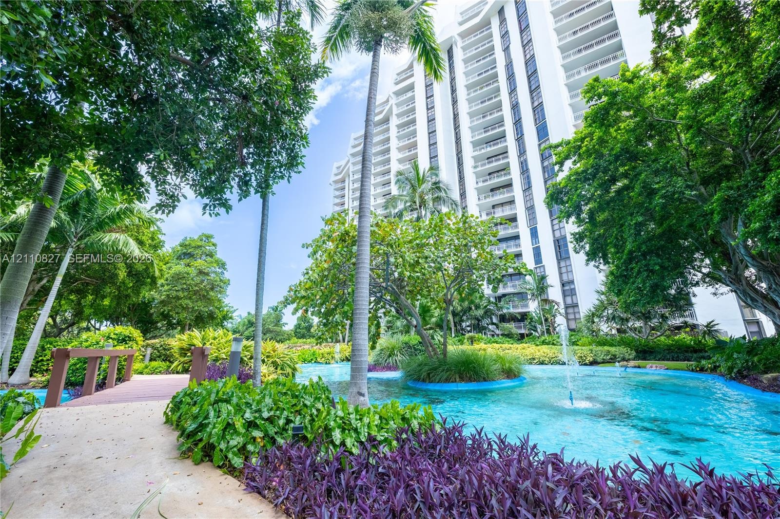 Multi-story high-rise building featuring a landscaped courtyard with a decorative pond, integrated fountains, mature palm trees, and a pedestrian foot