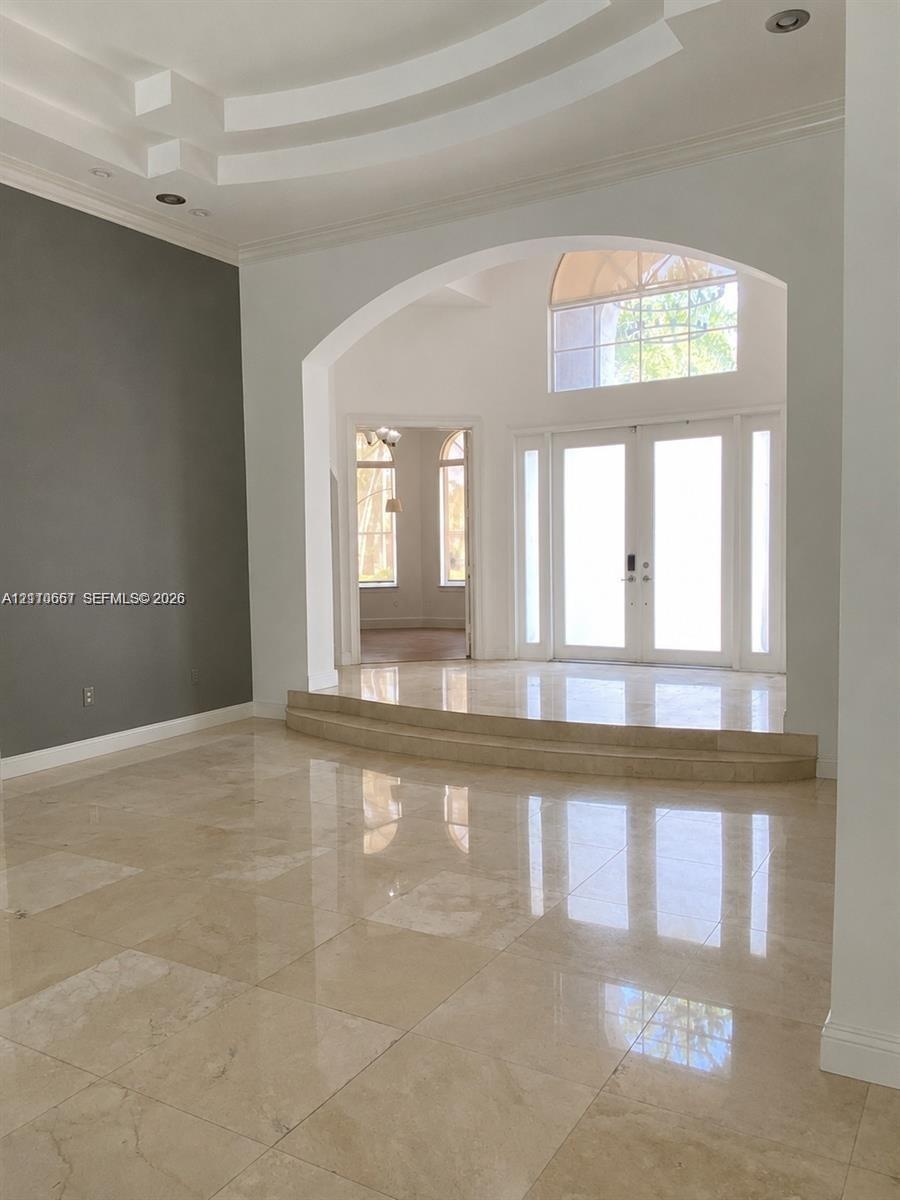 Grand foyer featuring polished stone flooring, a multi-tiered tray ceiling, an arched entryway, and double front doors with an overhead arched window