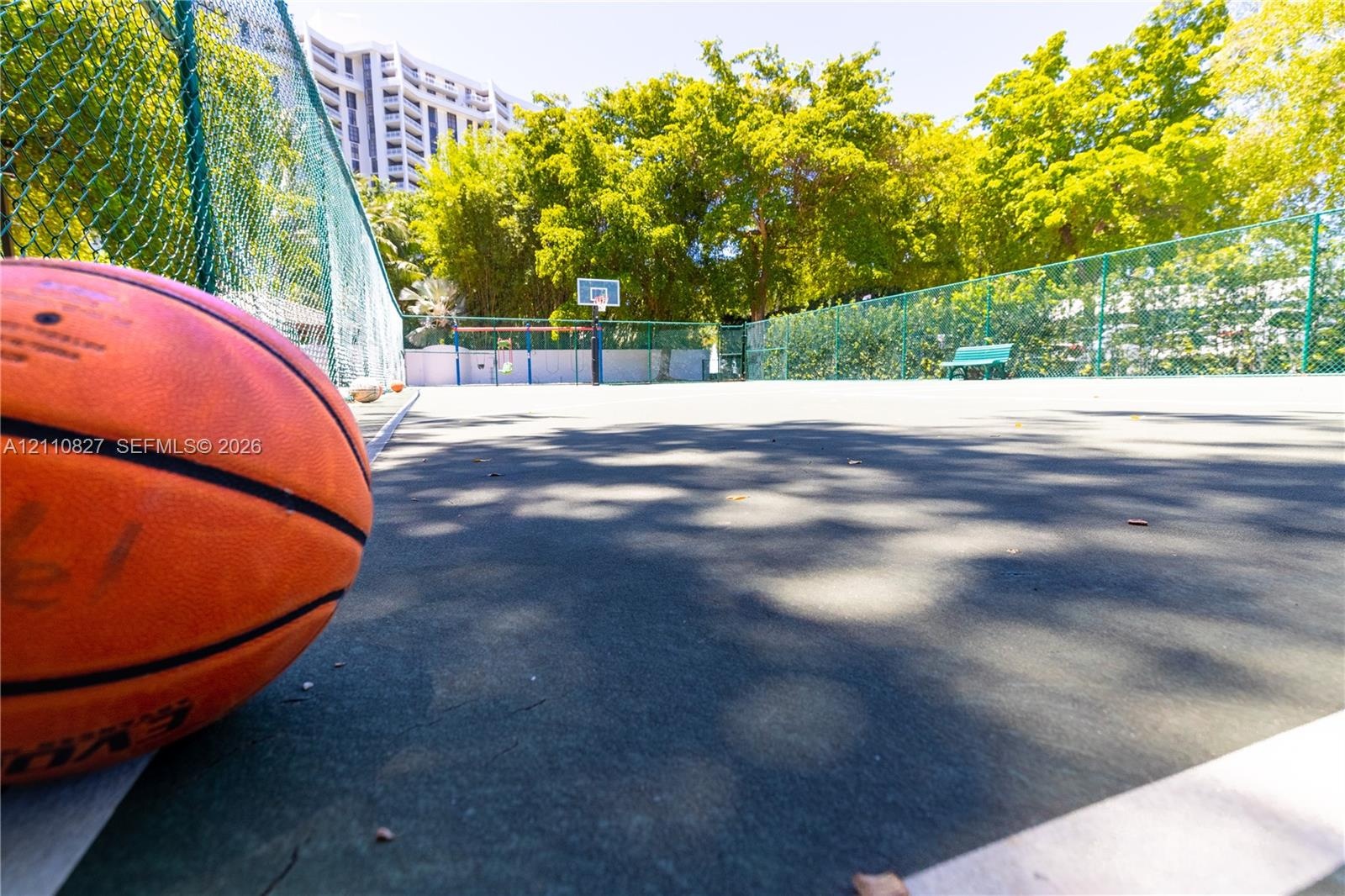 Recreational court featuring a hard surface, perimeter chain-link fencing, a basketball hoop, and a fixed bench