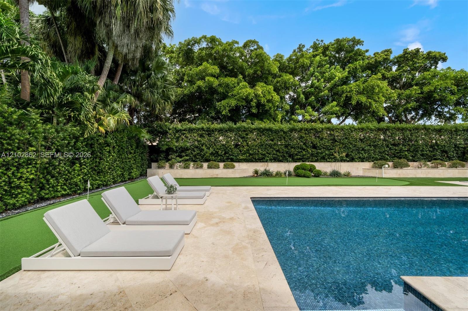 Private outdoor space featuring a rectangular swimming pool with stone-tiled decking, a manicured artificial turf putting green, and perimeter hedges