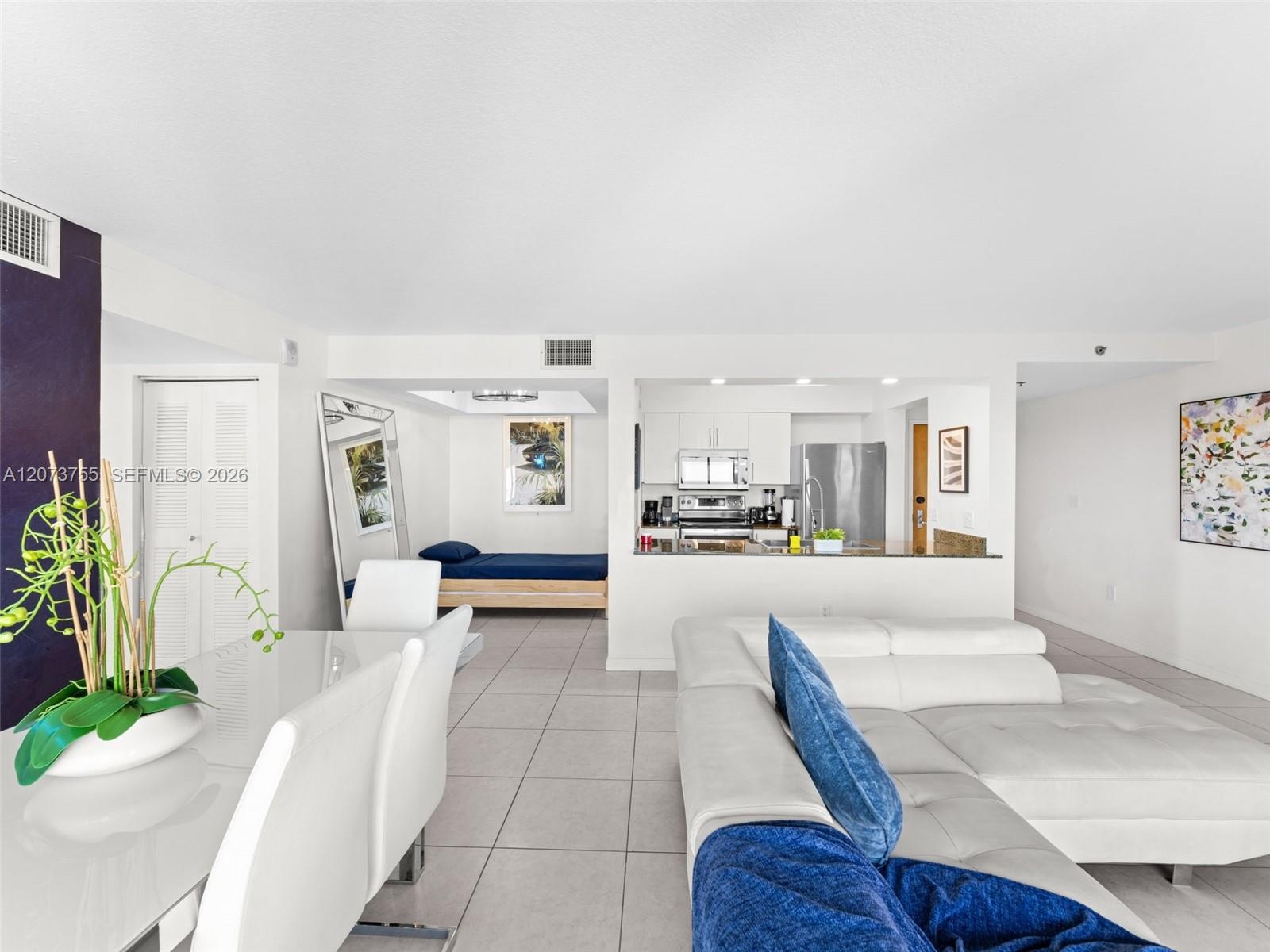 Open-concept interior featuring large-format tile flooring, kitchen with stainless steel appliances, neutral cabinetry, and a breakfast bar