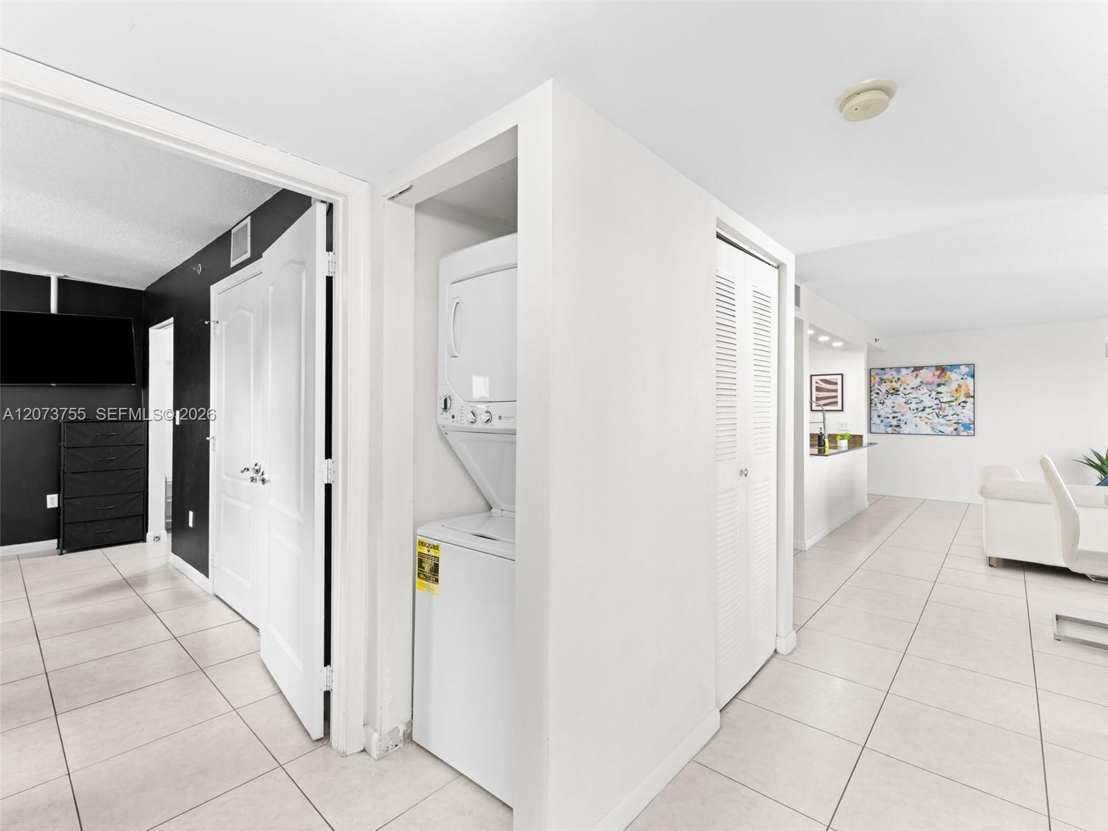 Dedicated laundry alcove with a stacked washer and dryer unit, neutral ceramic tile flooring, and white louvered bifold doors