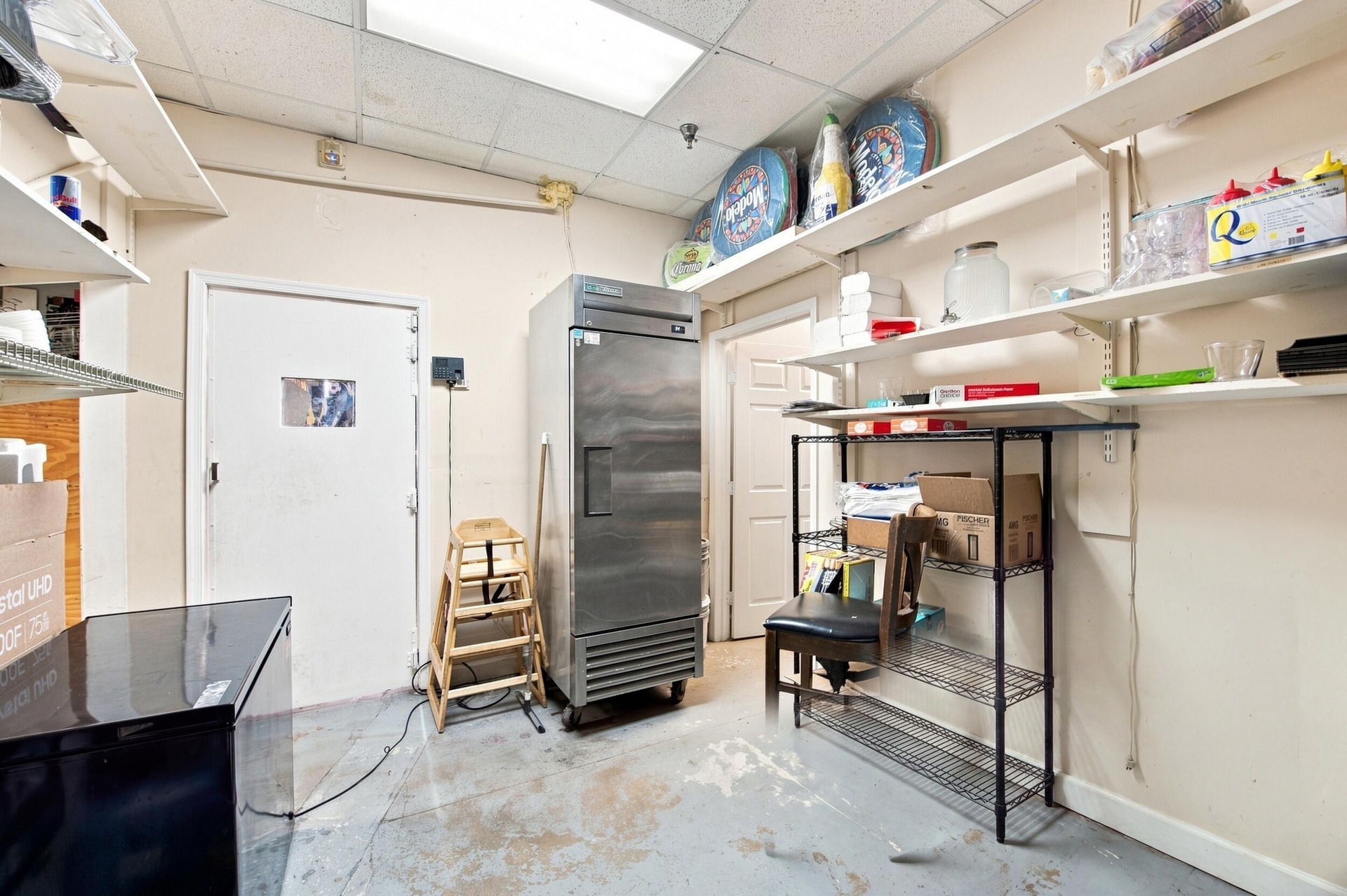 Storage area featuring wall-mounted adjustable shelving, a commercial-grade stainless steel refrigerator, drop ceiling with recessed lighting, and pai