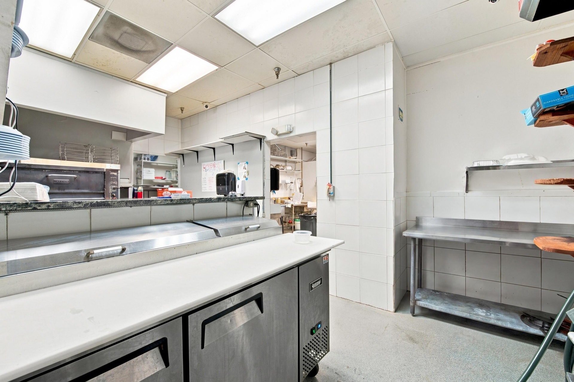 Commercial kitchen space featuring stainless steel refrigerated under-counter units, solid surface prep counters, wall-mounted stainless steel shelvin