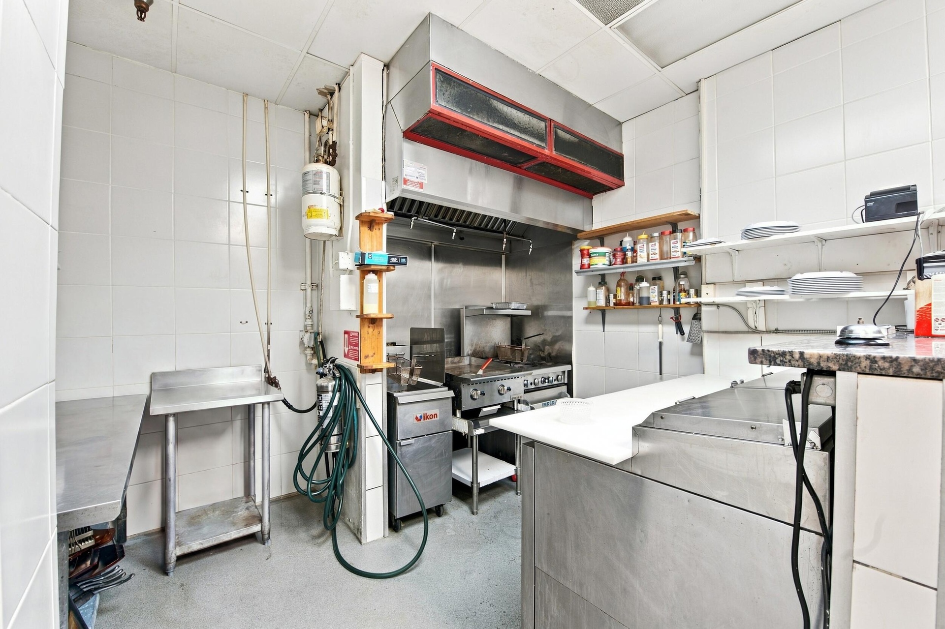 Commercial kitchen space featuring stainless steel preparation counters, a heavy-duty exhaust hood, industrial gas range, and wall-mounted shelving