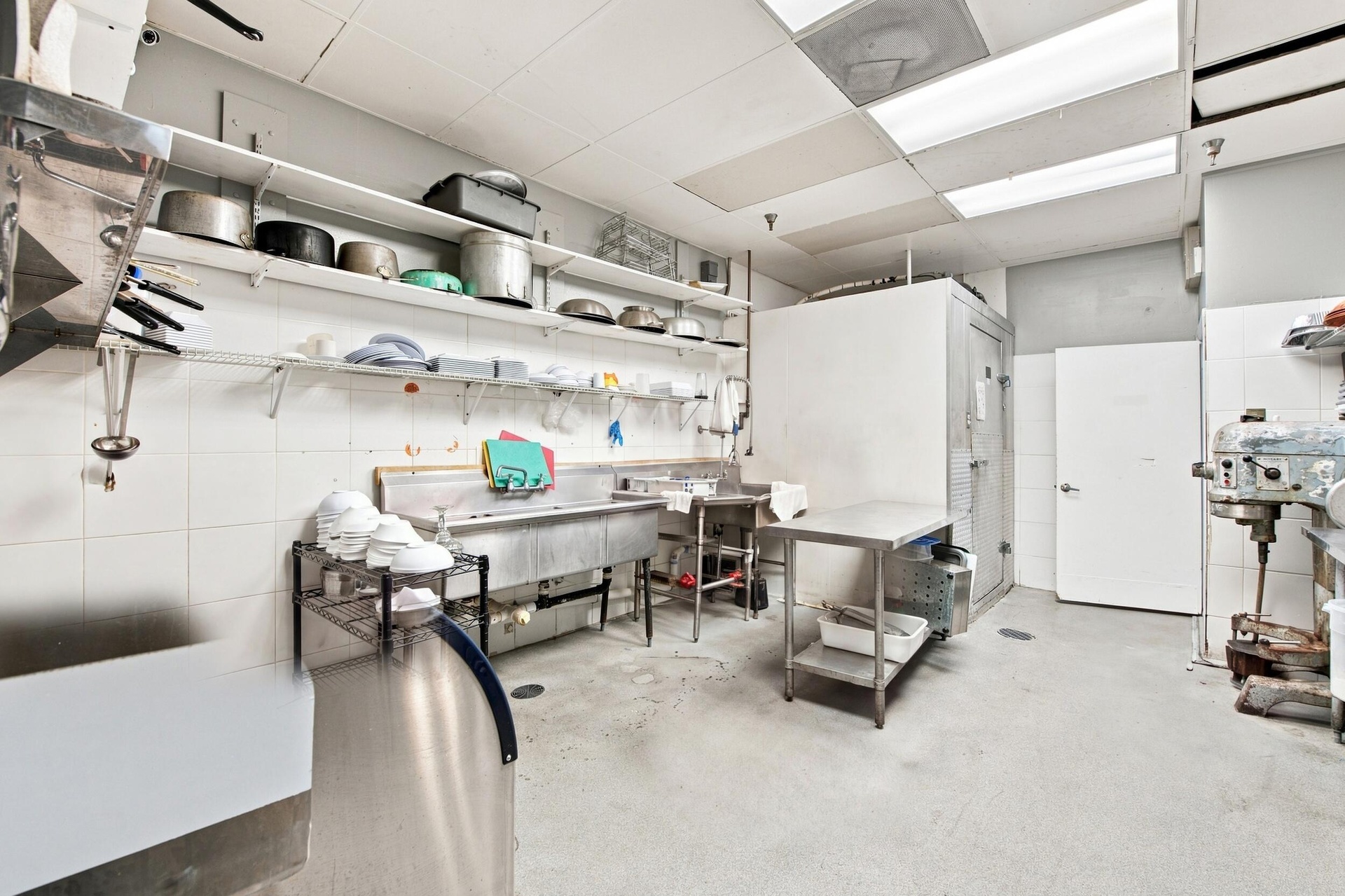 Commercial kitchen featuring stainless steel multi-basin sinks, wall-mounted open shelving, a walk-in cooler, and tiled wall surfaces