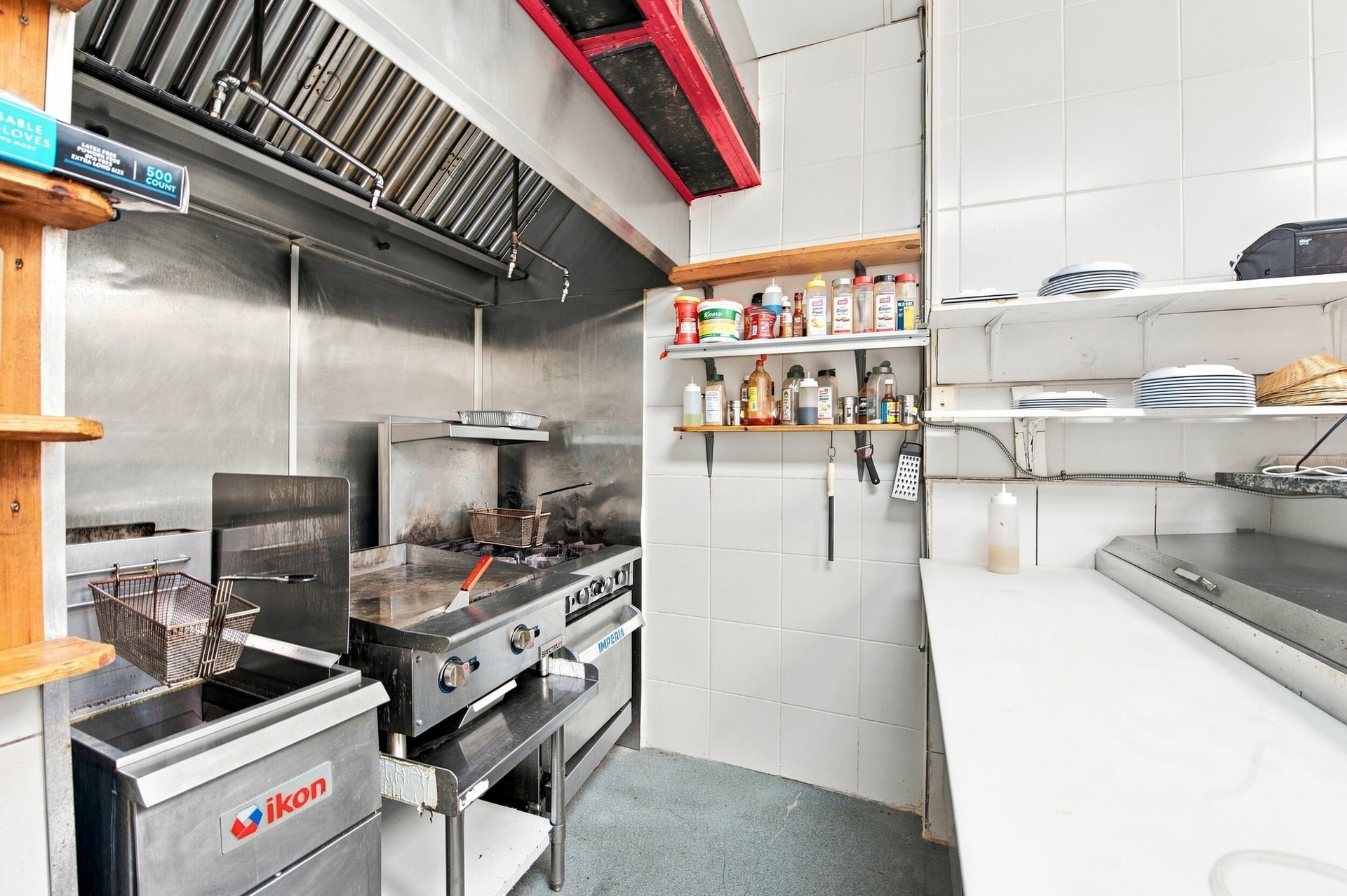 Commercial kitchen space featuring stainless steel wall cladding, professional-grade ventilation hood, built-in flat-top grill, deep fryer, and white 