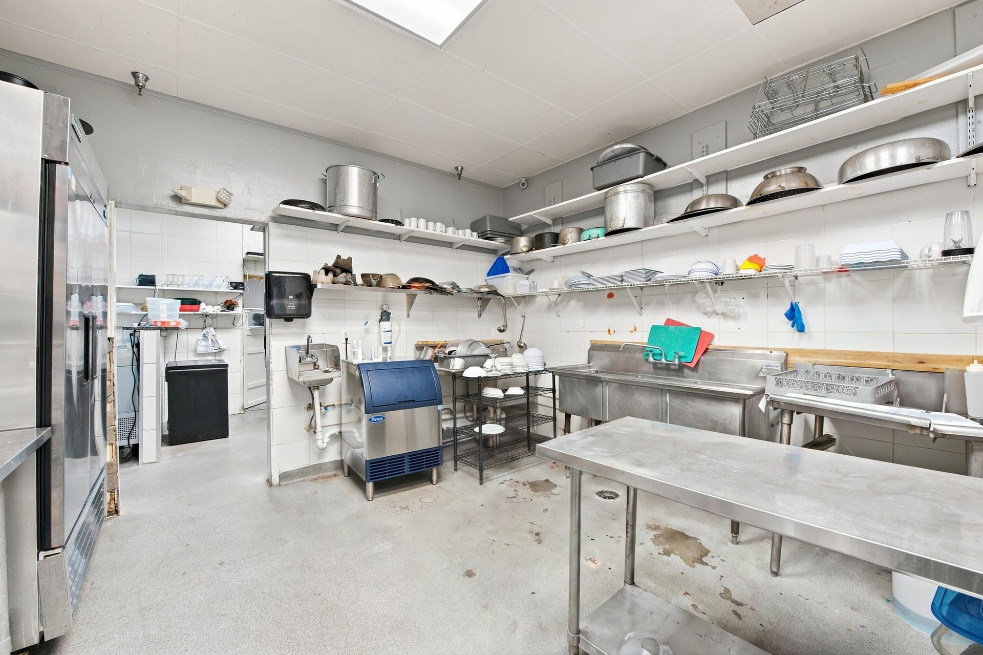 Commercial kitchen featuring stainless steel prep tables, multi-basin stainless steel sink, wall-mounted wire shelving, and industrial-grade flooring