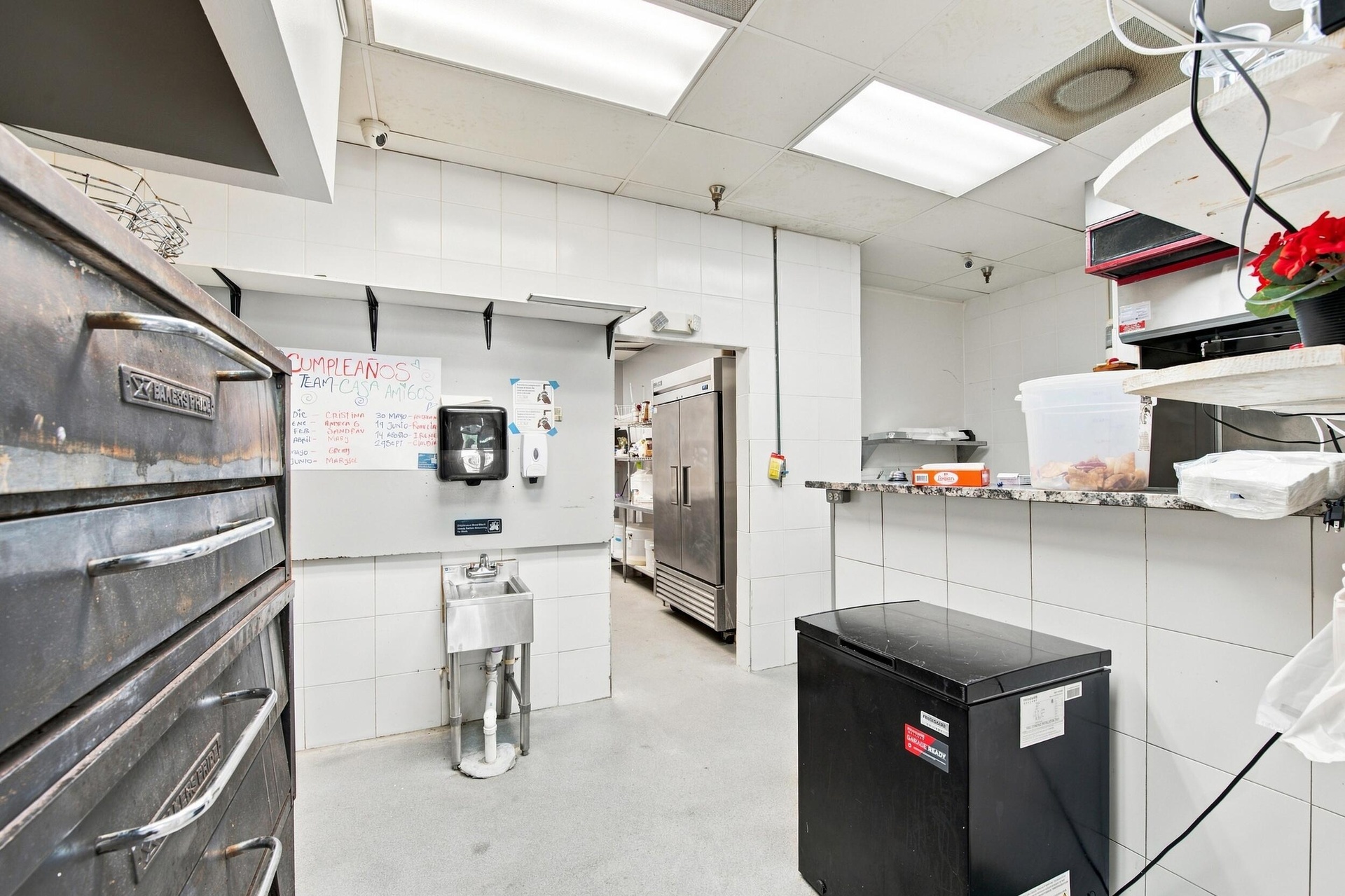 Commercial kitchen workspace featuring tiled wall finishes, a stainless steel basin, dual-door refrigeration unit, and a stone-surface service counter