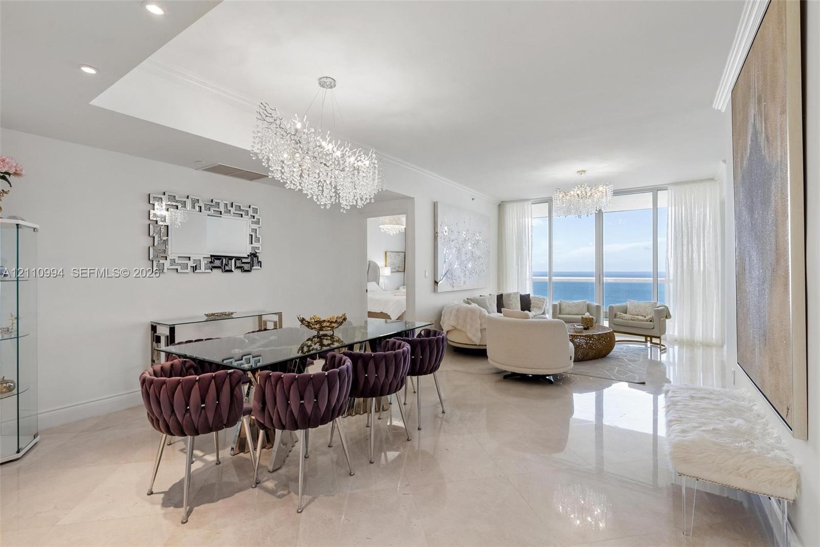 Open-concept interior featuring light-toned polished stone flooring, recessed ceiling lighting, statement crystal chandeliers, and expansive floor-to-