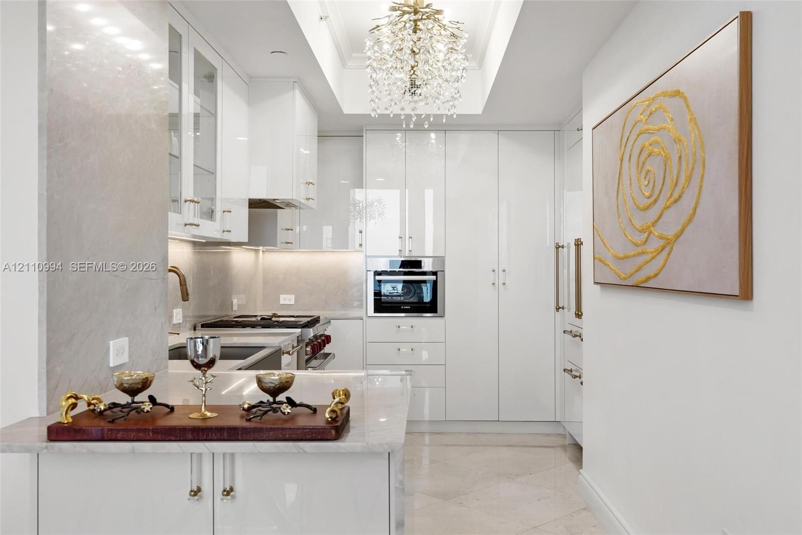 High-gloss white cabinetry featuring polished hardware, stone-finish countertops, recessed tray ceiling with crystal-style chandelier, and built-in st