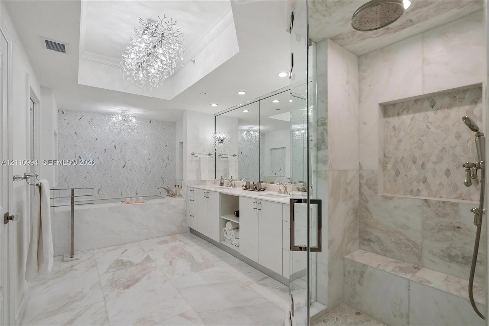 Bathroom featuring a white double-vanity, walk-in glass-enclosed shower with a built-in bench, soaking tub with decorative tile surround, and crystal 