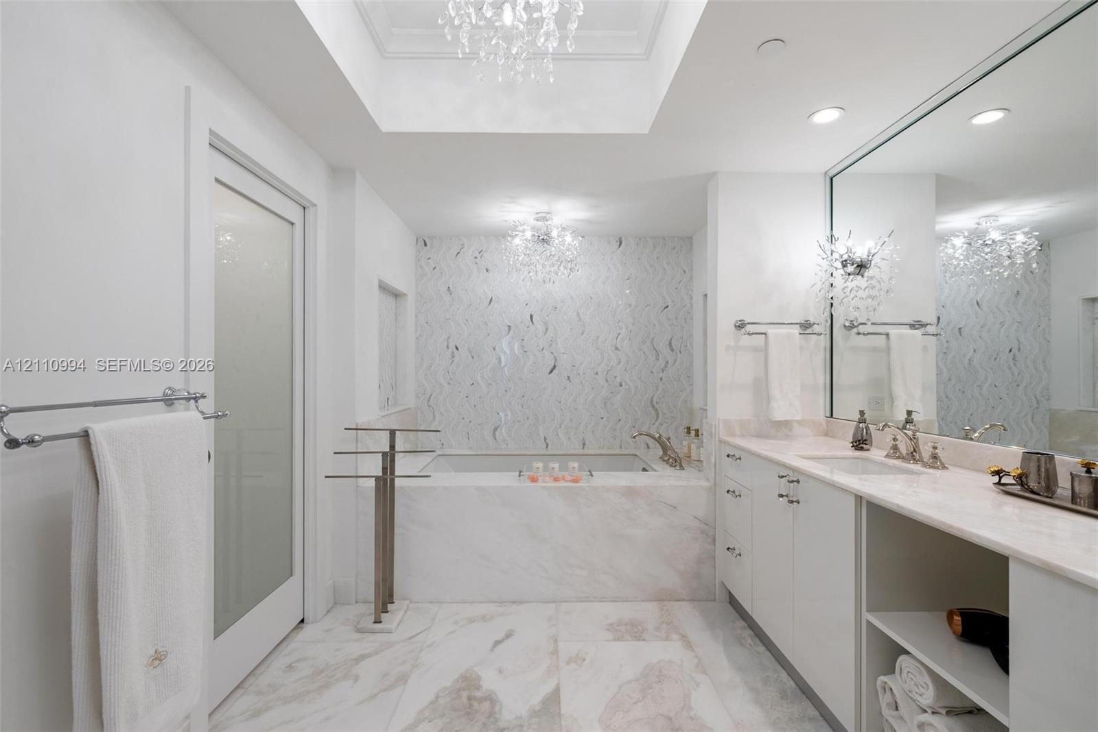 Soaking tub with stone surround, custom tiled accent wall, skylight with recessed tray ceiling, dual crystal chandeliers, and white cabinetry with sto