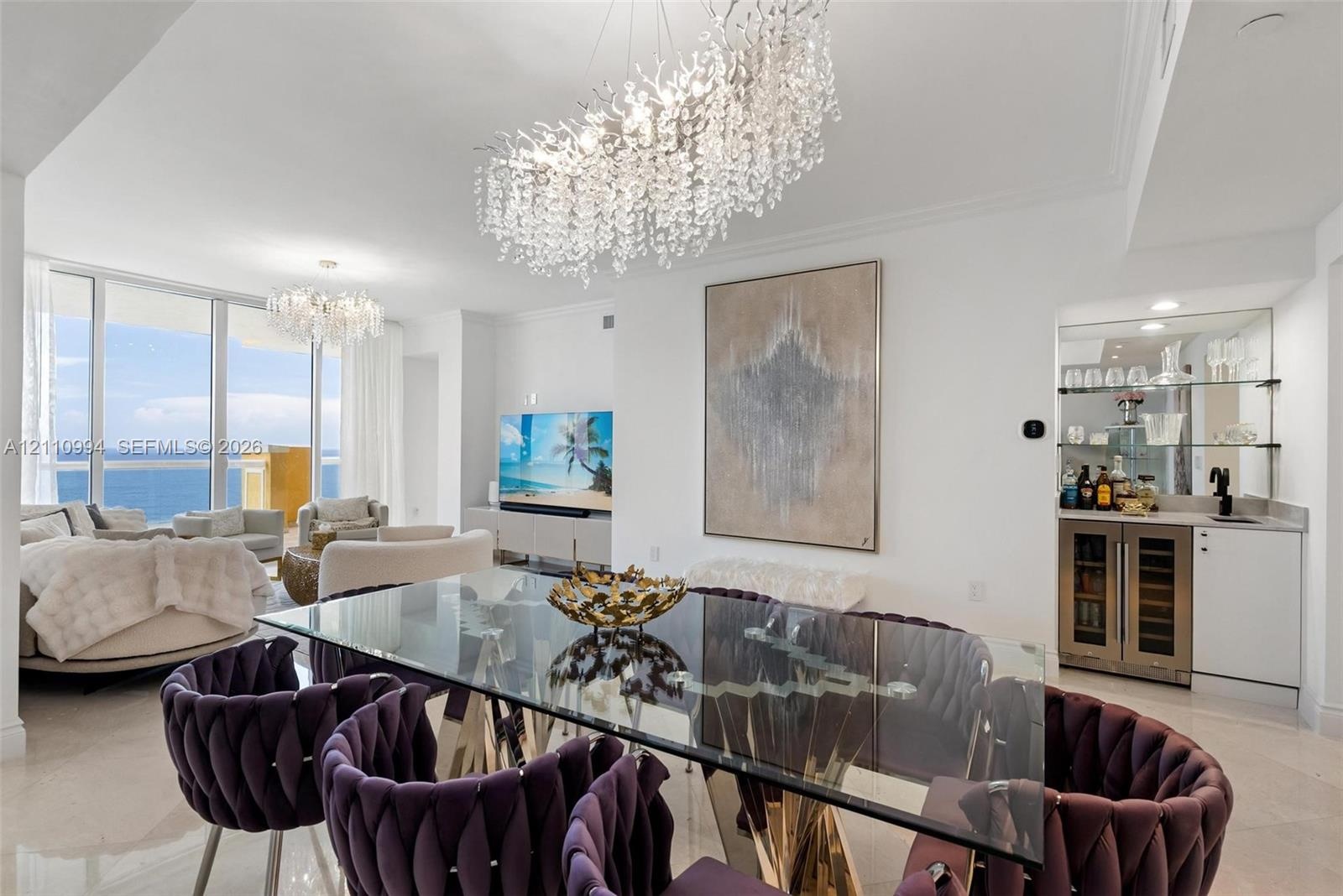 Open-concept space featuring floor-to-ceiling glass, custom crystal chandeliers, polished stone flooring, and a built-in wet bar with a glass-front be