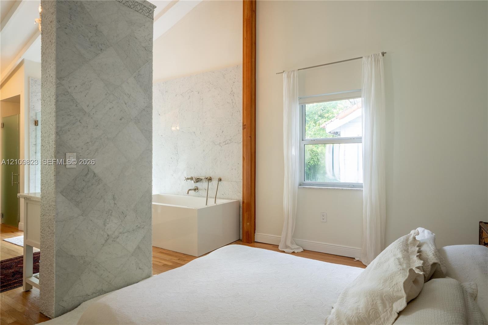 Vaulted ceiling, stone-tiled privacy wall, deep-soaking tub with wall-mounted plumbing, natural wood-finish flooring, and exposed vertical wood struct