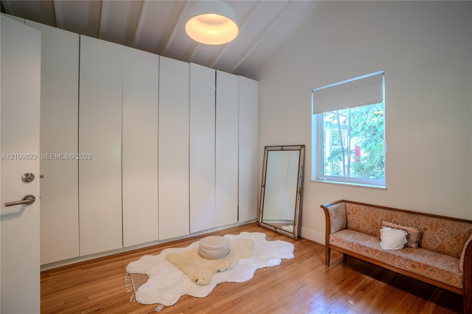 Floor-to-ceiling built-in cabinetry, vaulted ceiling with exposed beams, wood-finish flooring, and single-pane window with roller shade