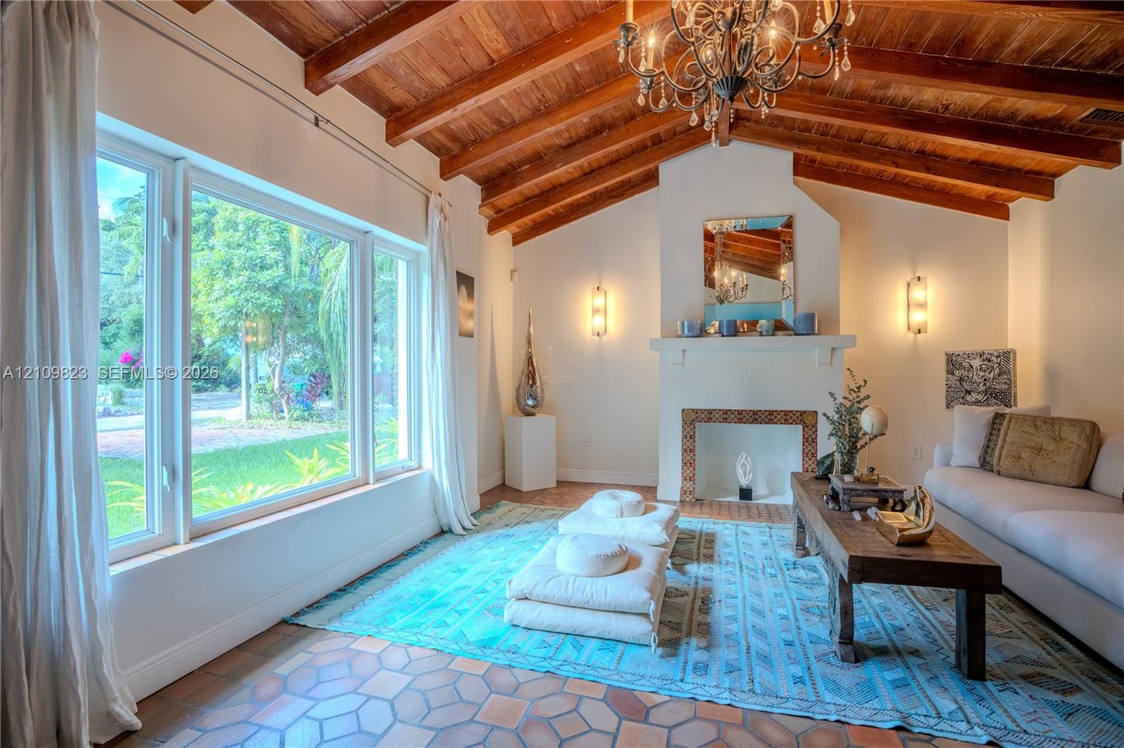 Vaulted ceiling with exposed wood beams, fireplace featuring decorative tile surround, terracotta tile flooring, and oversized windows