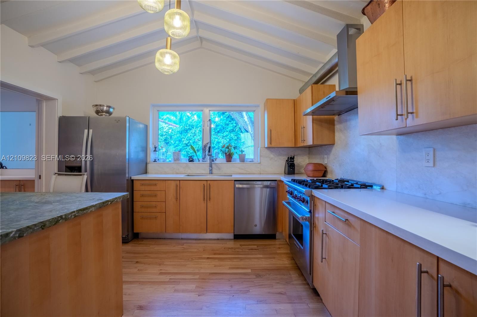 Kitchen featuring vaulted ceilings with exposed beams, light wood-finish cabinetry, stainless steel appliances, and a gas range with a chimney-style v