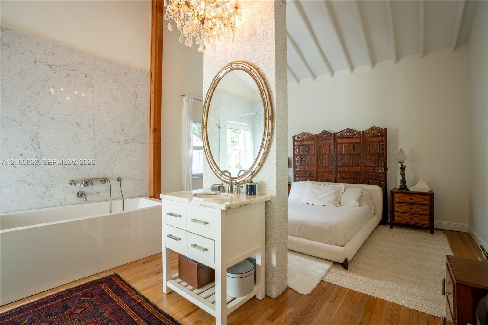Soaking tub with stone-tiled wall, white vanity with stone countertop, oval wall-mounted mirror, crystal chandelier, and exposed beam vaulted ceiling