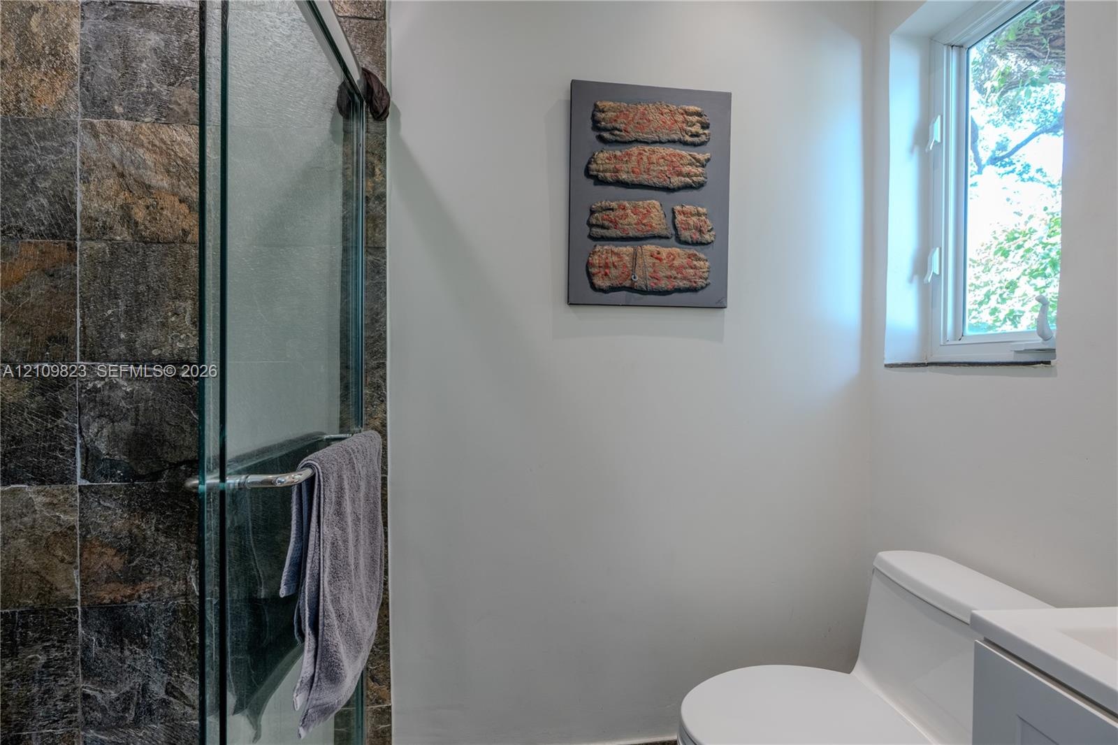 Shower enclosure featuring stone-textured wall tile and a glass door, paired with a white porcelain toilet and a single-pane window
