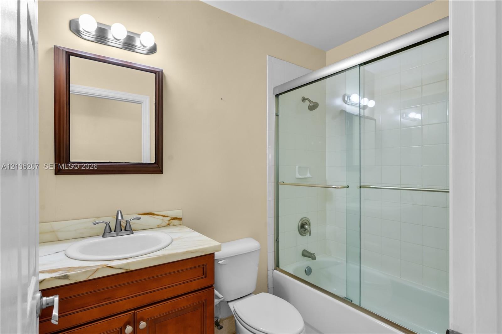 Bathroom featuring a vanity with a stone-top surface, oval basin, sliding glass tub-shower enclosure, wall-mounted mirror, and triple-bulb light fixtu