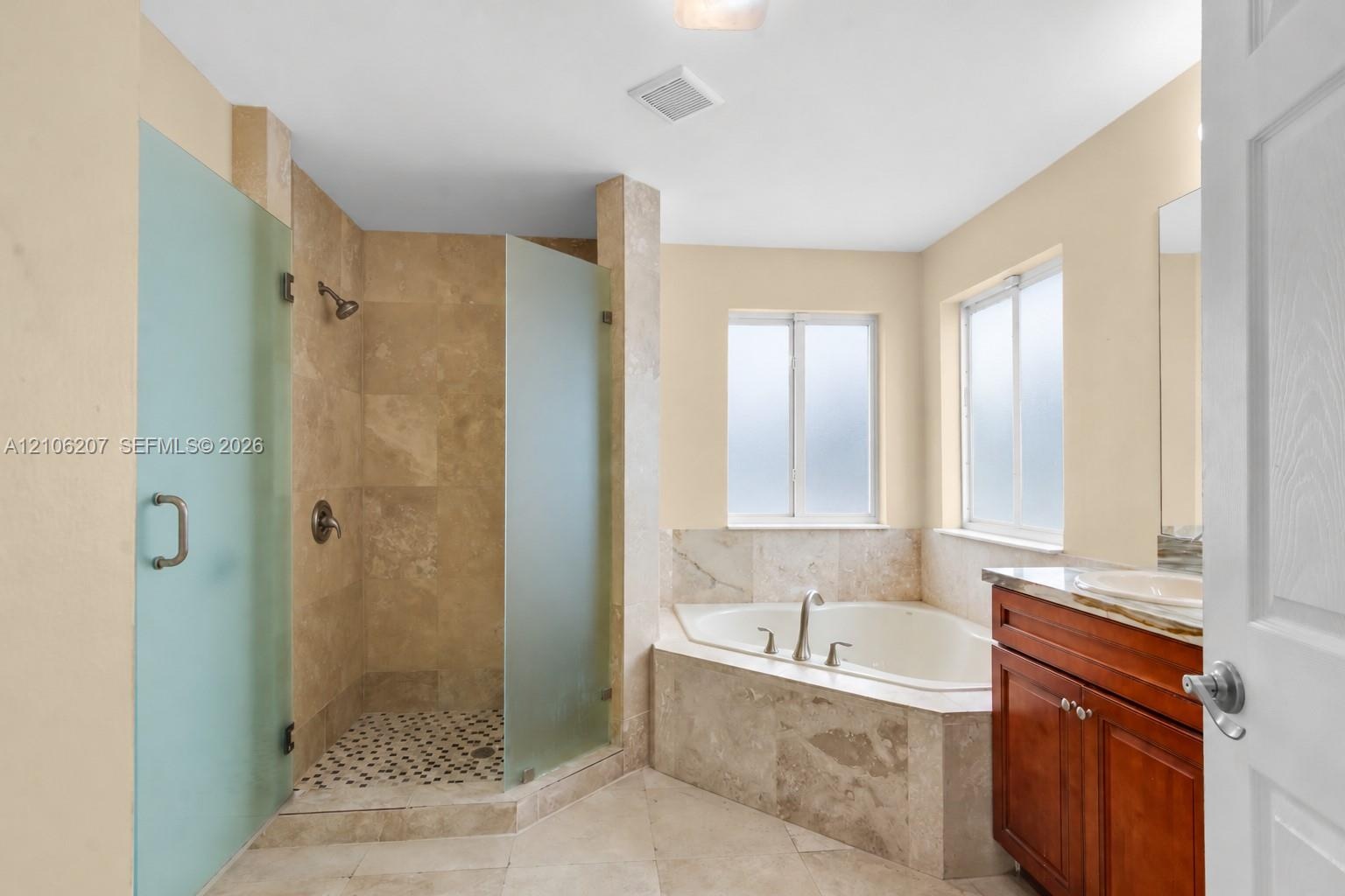 Bathroom featuring a corner soaking tub, walk-in shower with frosted glass enclosure, stone-tile surround, vanity with stone countertop, and an integr