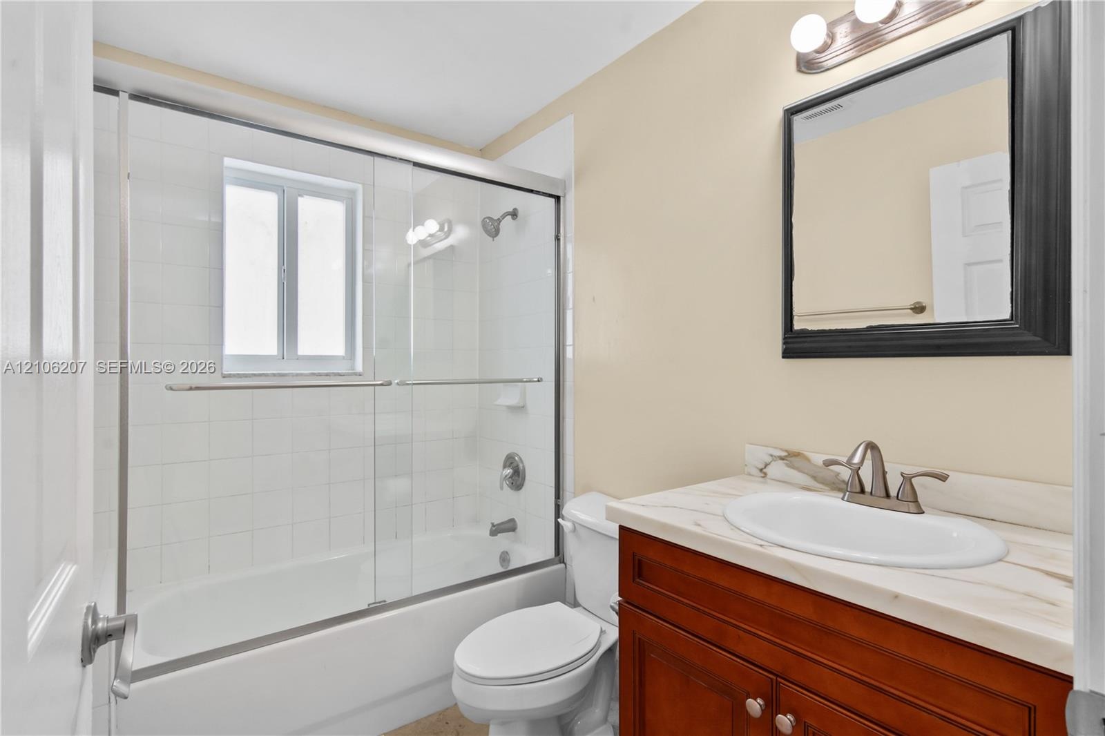 Bathroom featuring a wood-finish vanity with a stone-top integrated sink, a glass-enclosed shower and tub combination with tiled surround, and a wall-