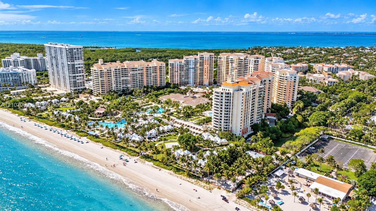 Aerial perspective of oceanfront high-rise structures, featuring expansive sandy coastline, turquoise water, manicured tropical landscaping, and outdo