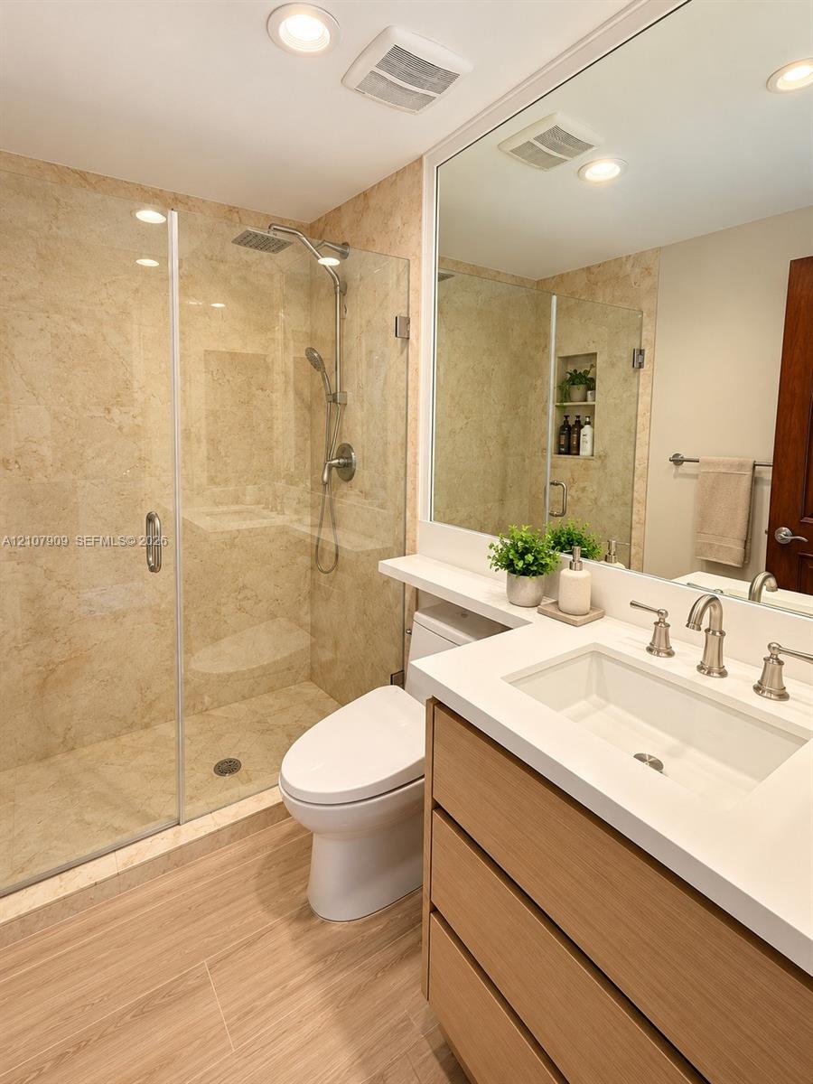 Bathroom featuring a frameless glass walk-in shower with stone wall tile, wood-finish cabinetry, quartz countertop with an integrated rectangular basi