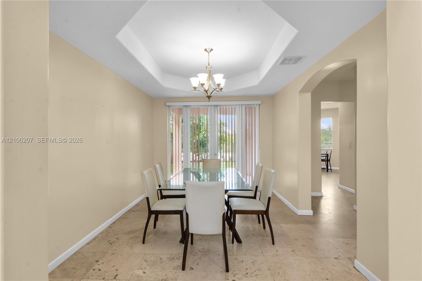 Tray ceiling with central chandelier, neutral wall tones, large-format stone-finish flooring, and glass doors with vertical blinds