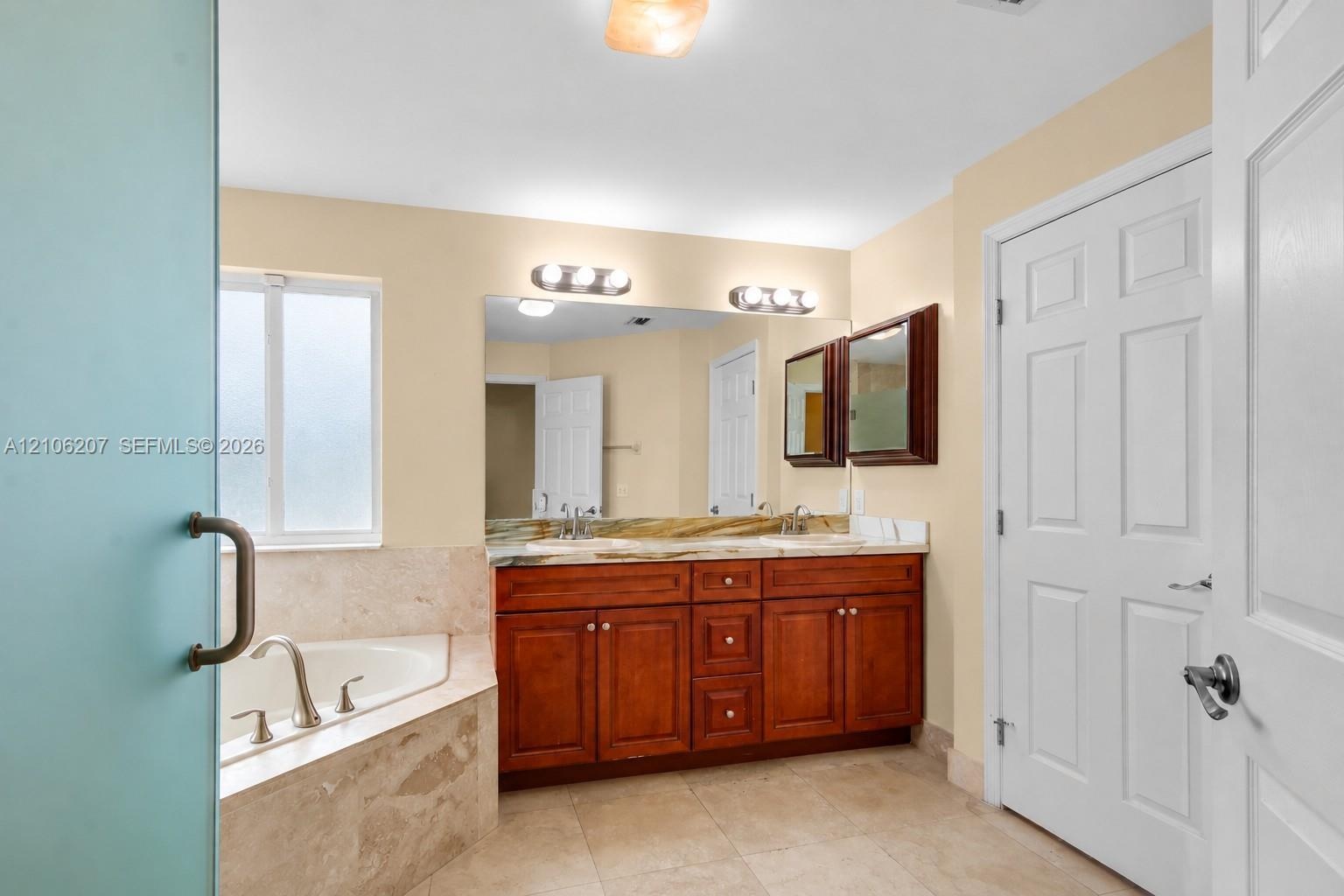 Bathroom featuring a double-sink vanity with wood cabinetry, stone-surround corner soaking tub, large wall-mounted mirror, and tile flooring