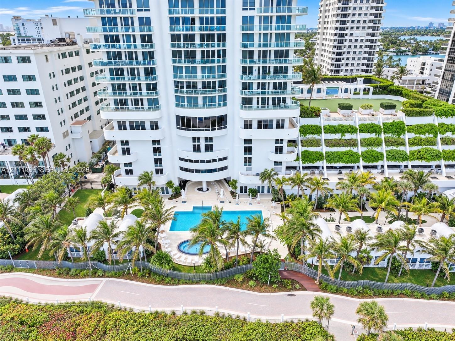 High-rise residential exterior featuring expansive glass balconies, a ground-level swimming pool, a separate spa, landscaped grounds with mature palm 