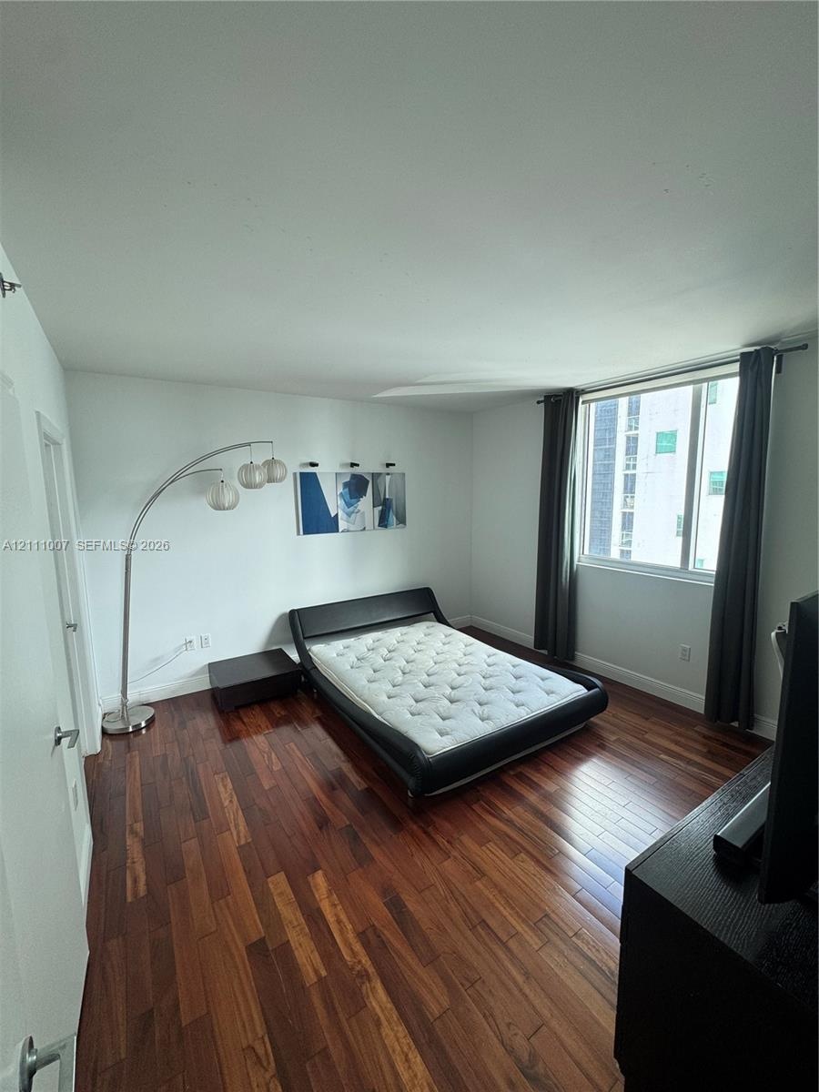 Bedroom featuring dark wood-finish flooring and a window with neutral-toned curtains