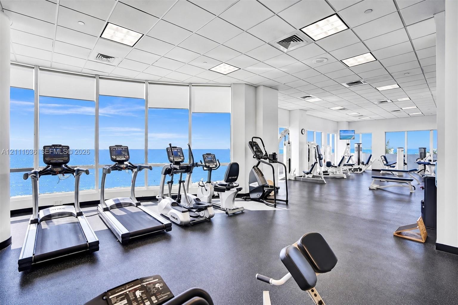 Fitness center featuring expansive floor-to-ceiling windows, panoramic ocean scenery, durable rubber flooring, and a suspended drop-tile ceiling