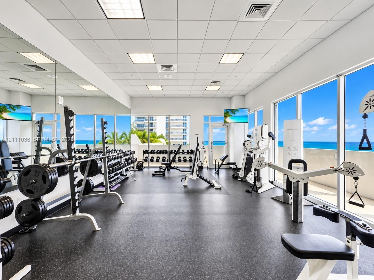 Fitness center featuring floor-to-ceiling windows, full-wall mirrors, rubberized flooring, and drop-ceiling tiles