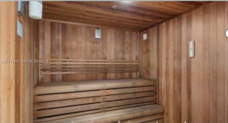 Sauna featuring wood paneling, multi-level slatted wooden benches, and wall-mounted lighting fixtures