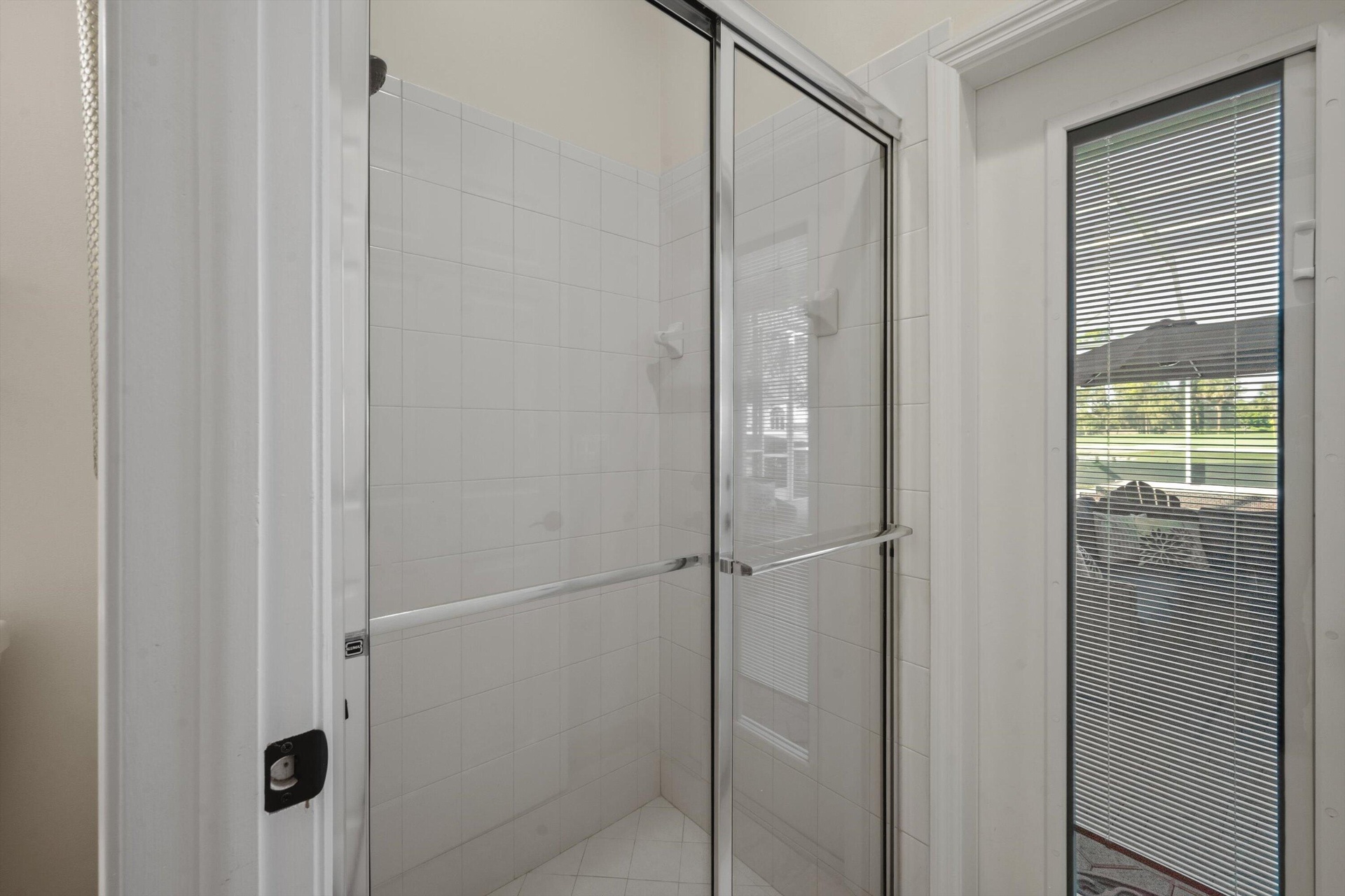 Shower enclosure featuring a chrome-framed sliding glass door, white tiled walls, and an exterior door with integrated blinds