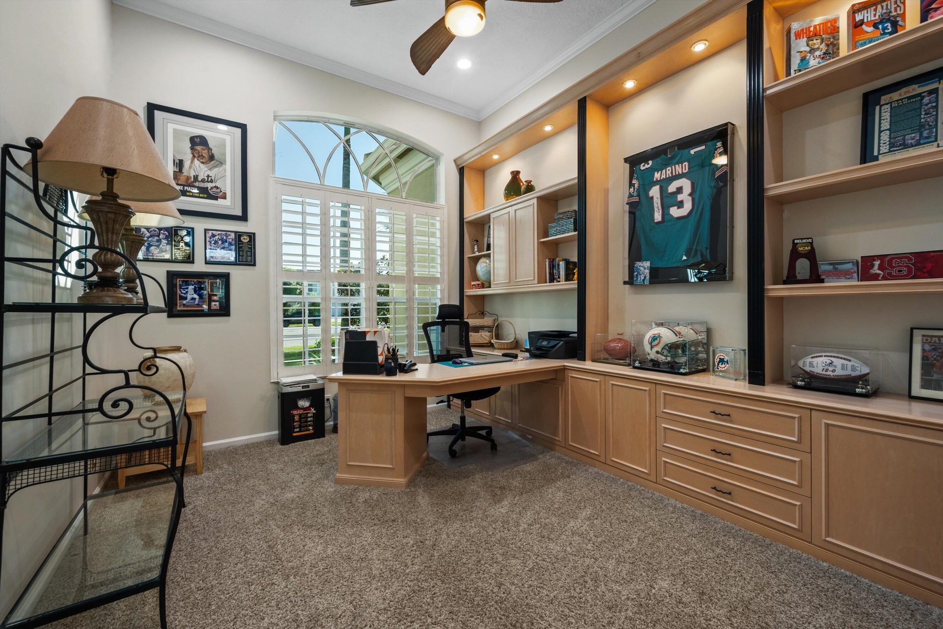Built-in light-wood cabinetry with integrated shelving and recessed accent lighting, arched window with plantation shutters, and ceiling fan fixture