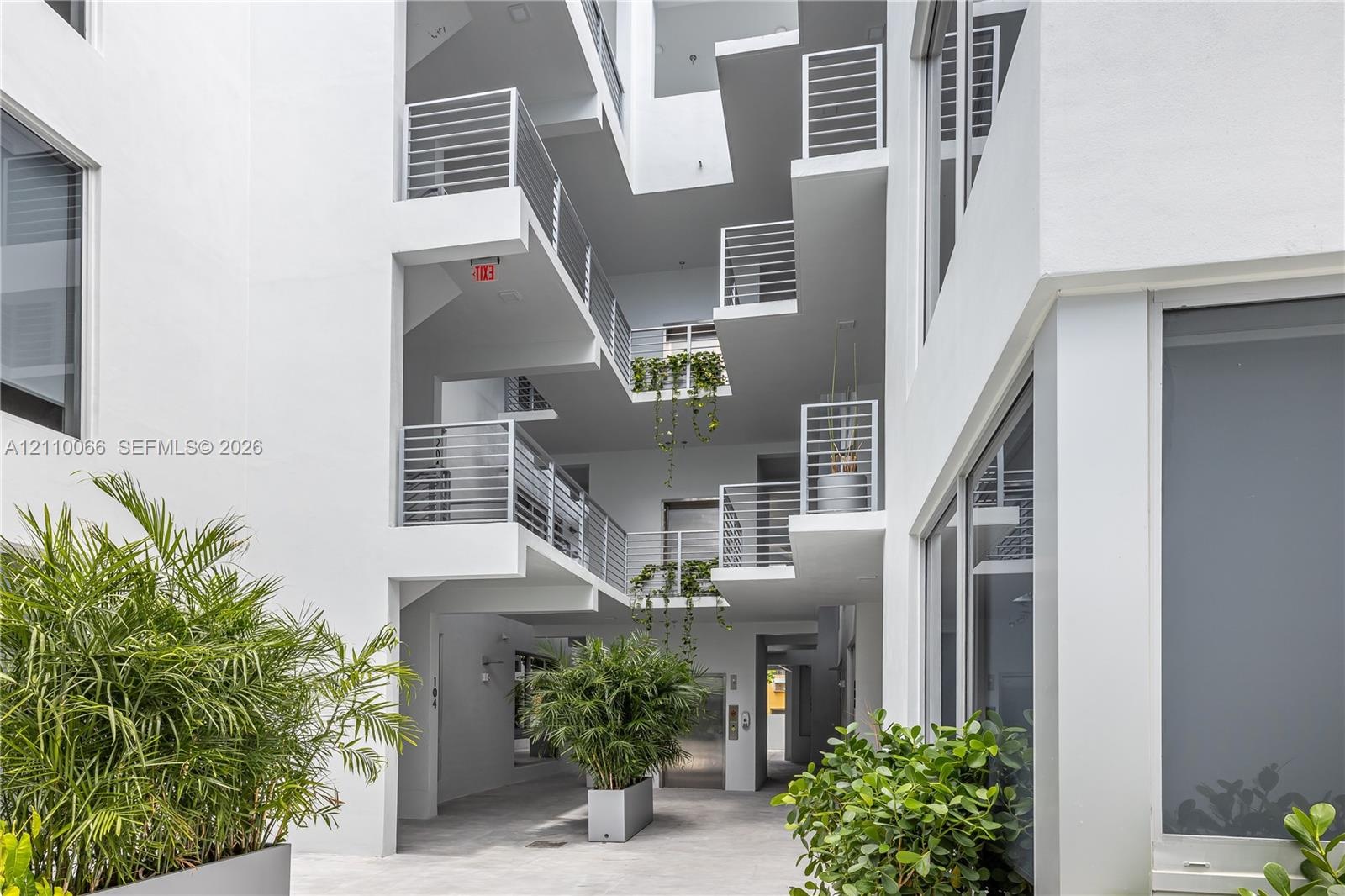 Multi-story architectural exterior featuring a white stucco facade, tiered balconies with horizontal metal railings, floor-to-ceiling glass windows, a