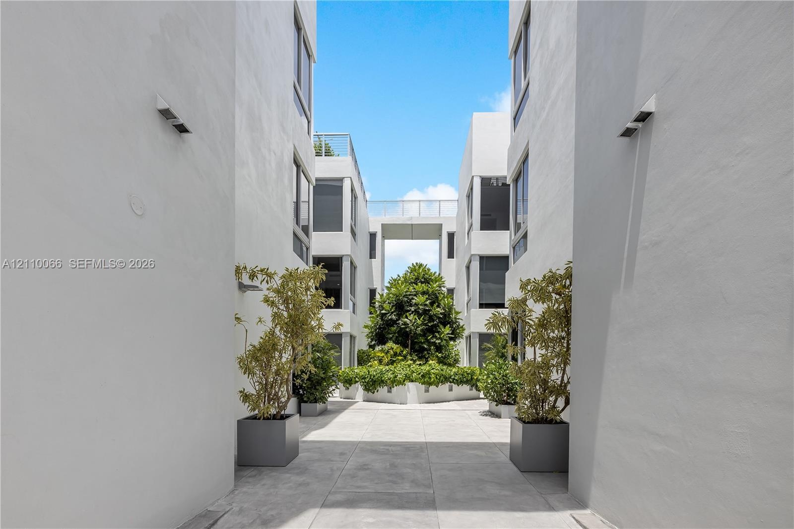 Modern multi-story exterior featuring smooth stucco siding, large windows, an overhead structural walkway, and central courtyard landscaping with ston