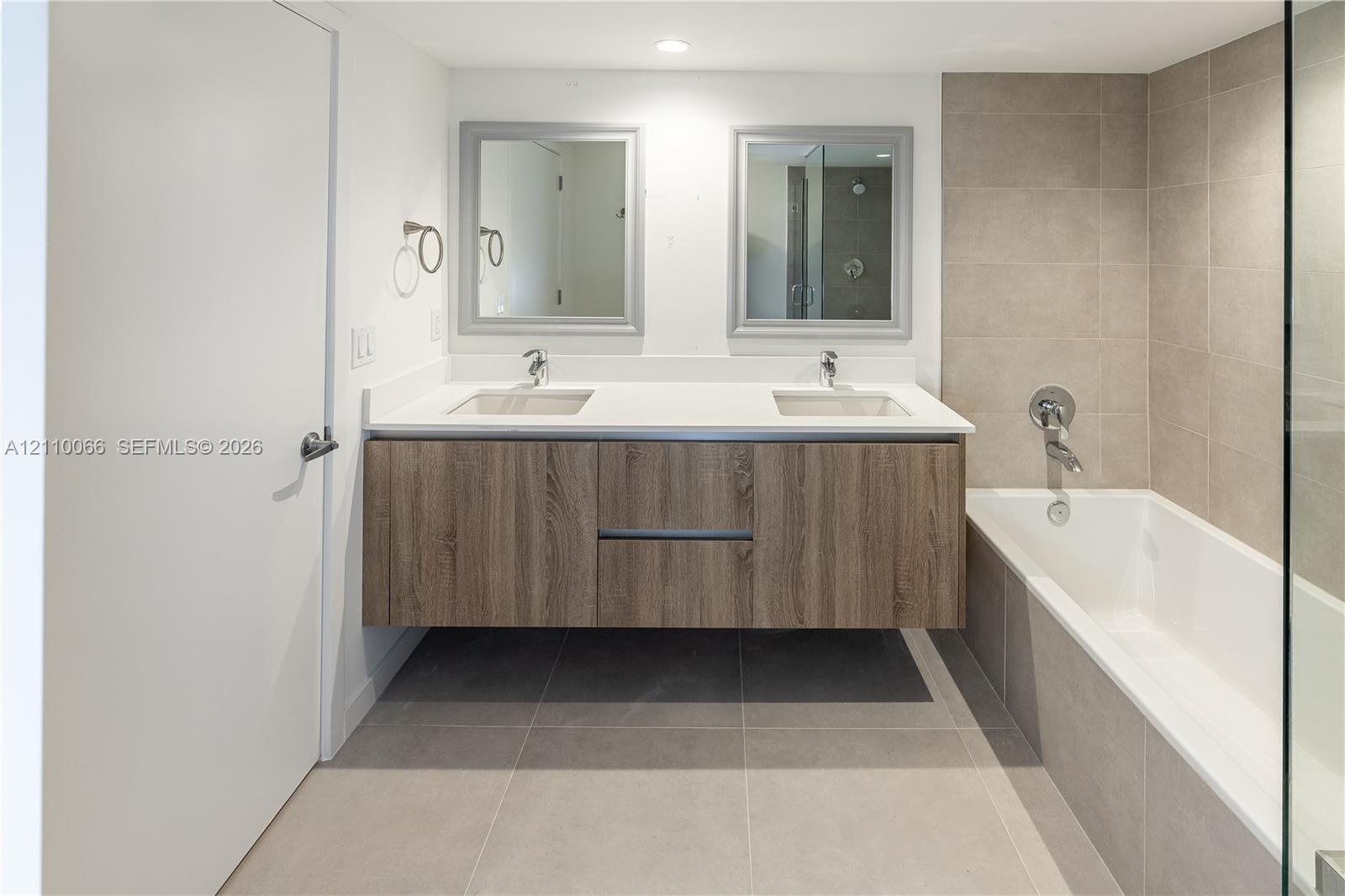 Floating dual-sink vanity with wood-finish cabinetry, solid surface countertop, large-format tile flooring, and integrated shower-tub combination feat