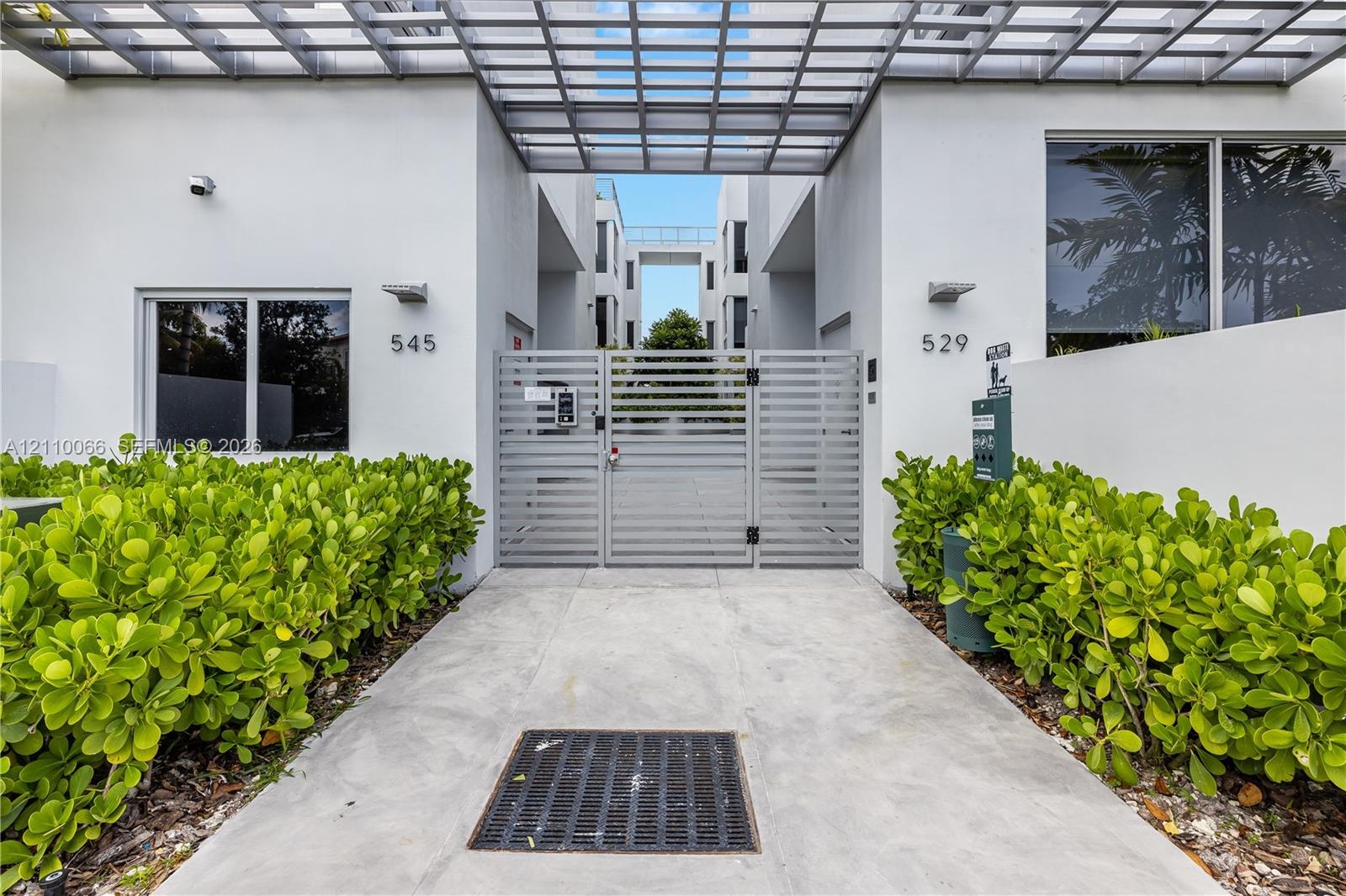 Modern exterior featuring smooth-stucco facade, horizontal slatted metal security gate, overhead metal trellis, and integrated wall-mounted lighting