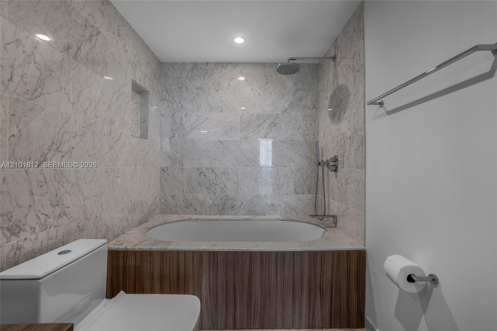 Full-height stone wall tiling, built-in soaking tub with wood-finish paneling, recessed shower niche, ceiling-mounted rainfall showerhead, and wall-mo